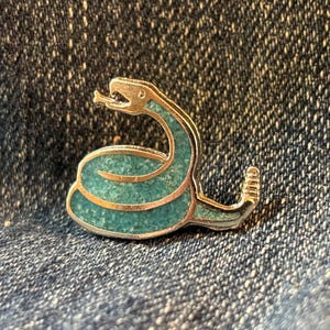 May include: A silver-toned snake pin with a turquoise-colored body. The snake is coiled, with its head raised and tongue extended. The body of the snake is filled with small, turquoise-colored stones, and the pin has a detailed design.