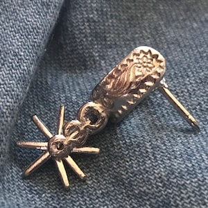 May include: Silver cowboy spur earring with a star-shaped design and a detailed floral pattern on the back.