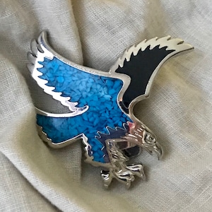 May include: A silver eagle brooch with blue and black inlay. The eagle is in flight with its wings spread wide. The inlay is made of small, turquoise-colored stones.