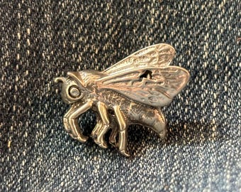 May include: A silver-colored bee pin with detailed wings, legs, and body. The bee pin is made of metal and is set against a textured denim background. The pin is a decorative accessory.