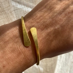 Könnte beinhalten: Ein goldfarbenes Armreif-Armband mit offenem Design. Das Armband hat eine glatte, matte Oberfläche und eine schlichte, elegante Form. Die Enden des Armbands sind leicht ausgestellt. Ein stilvolles Schmuckstück.