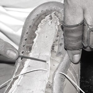 May include: A close-up of a person sewing a leather shoe with a needle and thread. The shoe is being stitched together with a white thread. The person is wearing a leather glove on their right hand.