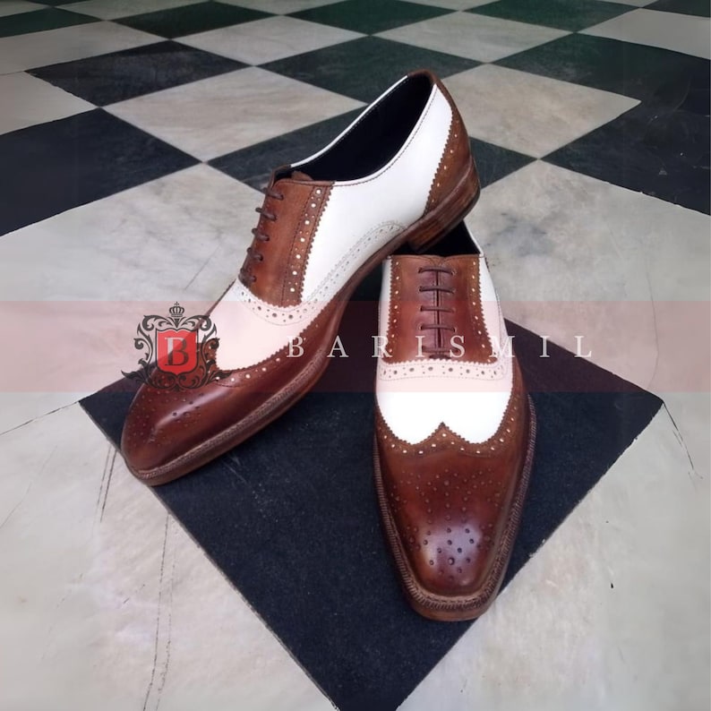 May include: A pair of brown and white leather Oxford shoes with wingtip detailing. The shoes have a lace-up closure and a brogue pattern.