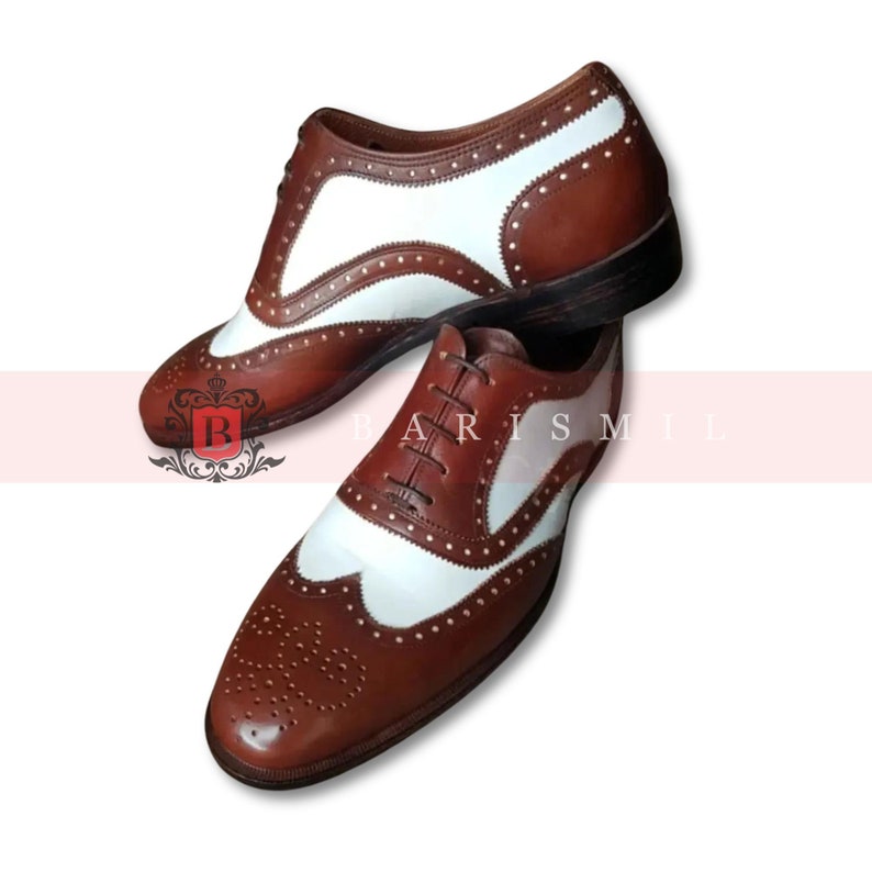 May include: Brown and white leather oxford shoes with wingtip detailing and a brogue pattern. The shoes have a lace-up closure and a rounded toe.