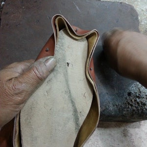 May include: A close-up of a brown leather shoe being worked on by a cobbler. The shoe is turned inside out, revealing the insole and stitching. The cobbler is using a tool to work on the shoe.