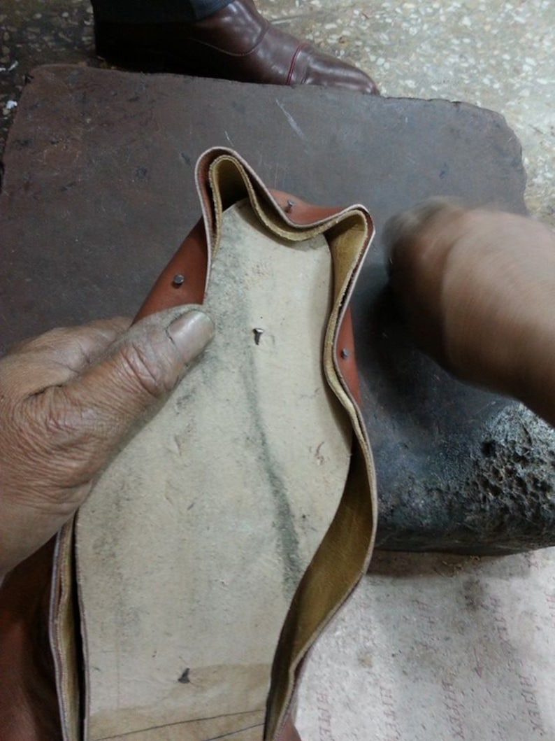 May include: A close-up of a brown leather shoe being worked on by a cobbler. The shoe is turned inside out, revealing the insole and stitching. The cobbler is using a tool to work on the shoe.