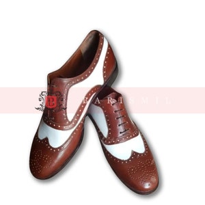 May include: Brown leather oxford shoes with white leather accents and a wingtip design. The shoes have a lace-up closure and a brown sole.