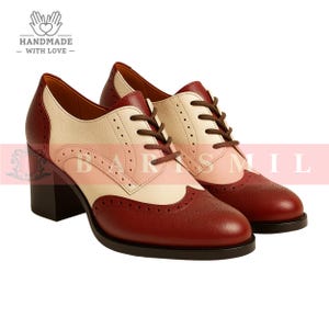 K&ouml;nnte beinhalten: Zweifarbige Oxford-Schuhe aus Leder mit Blockabsatz. Die Schuhe haben eine bordeauxfarbene Spitze und Ferse, einen cremefarbenen Schaft und einen rosa Mittelteil. Braune Schn&uuml;rsenkel und ein dekoratives Lochmuster sind sichtbar. Die Abs&auml;tze sind ca. 7,6 cm hoch.