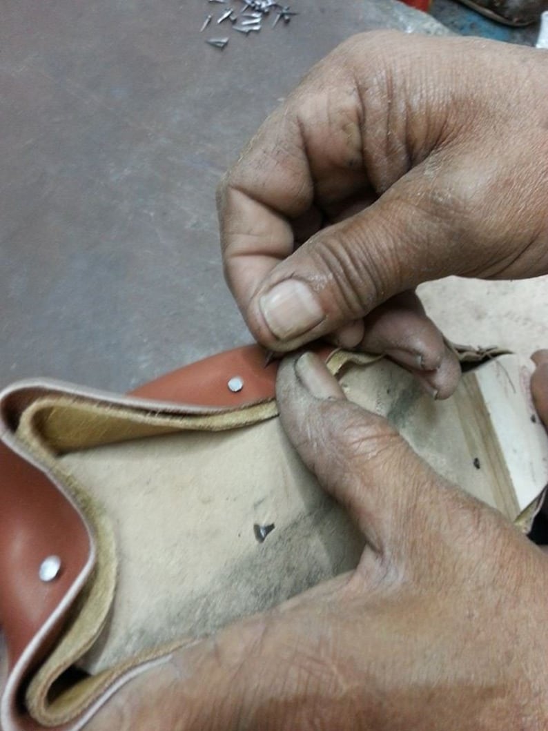 Puede incluir: Primer plano de un proceso de fabricaci&oacute;n de zapatos. Se ven manos trabajando en un zapato de cuero marr&oacute;n, asegurando el cuero con peque&ntilde;as tachuelas plateadas. El zapato tiene un interior de color beige claro.