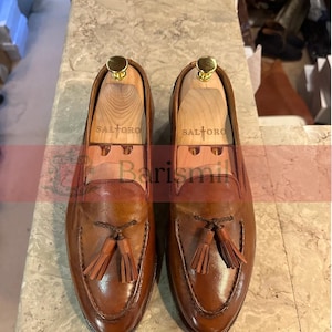 May include: A pair of brown leather tassel loafers with a stitched design. The loafers have a wooden shoe tree inside and the brand name "SALTORO" is printed on the shoe tree.