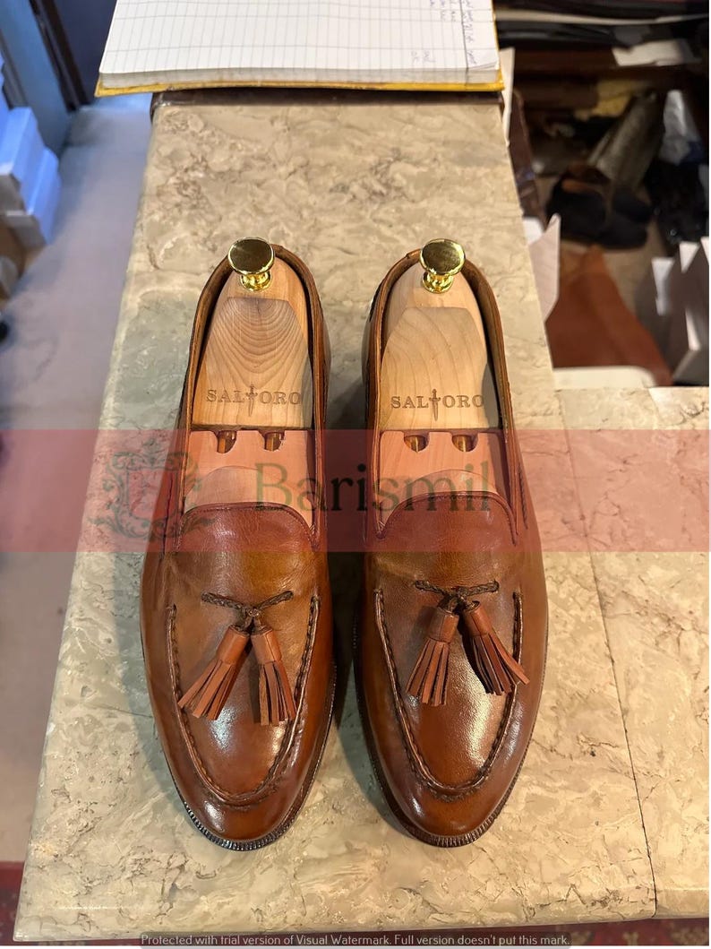 May include: A pair of brown leather tassel loafers with a stitched design. The loafers have a wooden shoe tree inside and the brand name "SALTORO" is printed on the shoe tree.