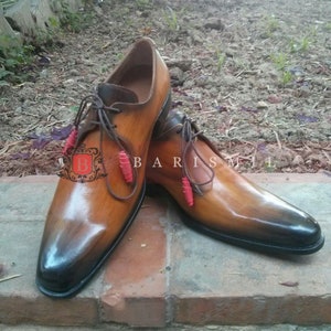 May include: A pair of brown leather lace-up shoes with a pointed toe. The shoes have a gradient color effect, transitioning from a darker brown to a lighter brown. The laces are brown and have red beads at the ends. The shoes are sitting on a brick surface.