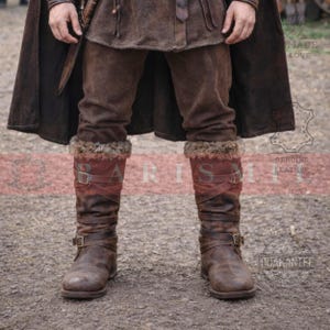 May include: Brown leather boots with fur trim and buckle straps. The boots are worn with brown pants and a dark brown cloak. The boots appear to be made of genuine leather.