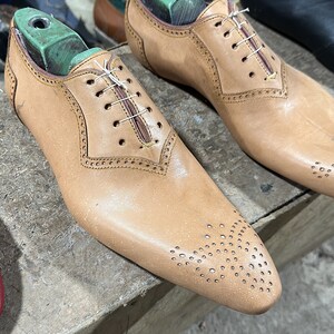 May include: Two brown leather shoes with laces and a perforated design on the toe. The shoes are on wooden shoe forms with green tops.