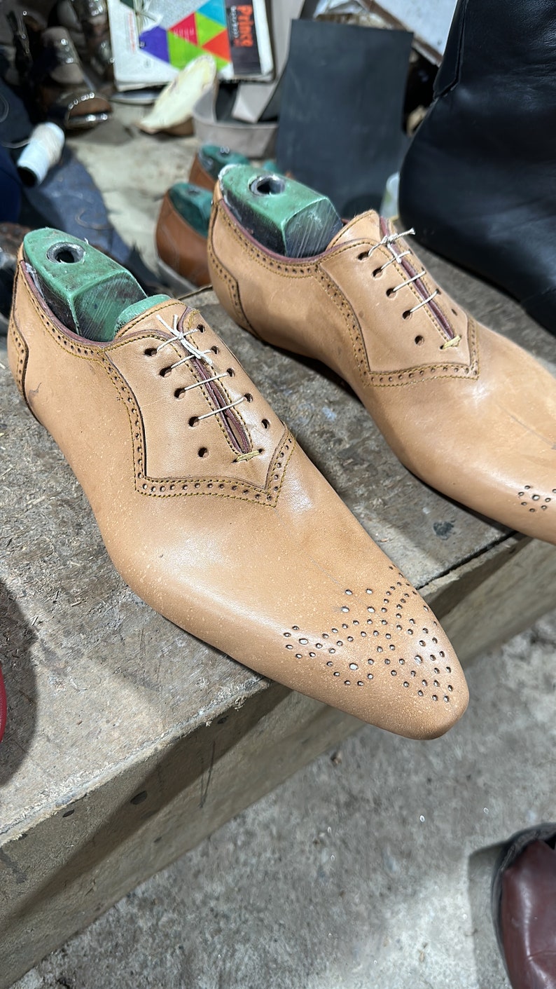 May include: Two brown leather shoes with laces and a perforated design on the toe. The shoes are on wooden shoe forms with green tops.