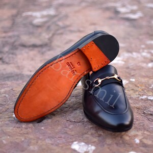 Op de afbeelding: Een paar zwarte leren loafers met een gouden gesp. De loafers hebben een bruine leren zool en zijn gemaakt in Itali&euml;.