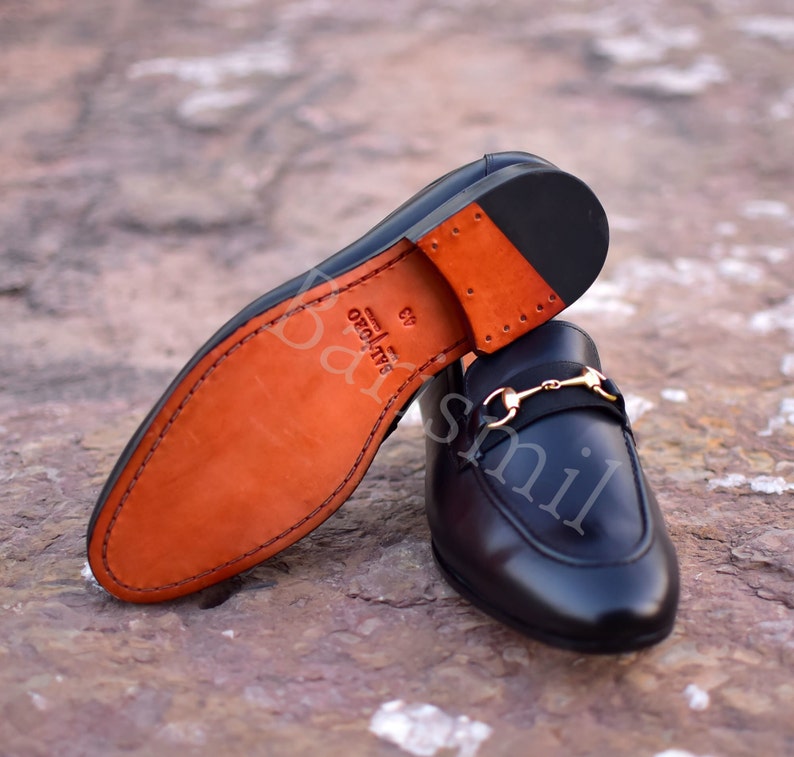 Op de afbeelding: Een paar zwarte leren loafers met een gouden gesp. De loafers hebben een bruine leren zool en zijn gemaakt in Itali&euml;.