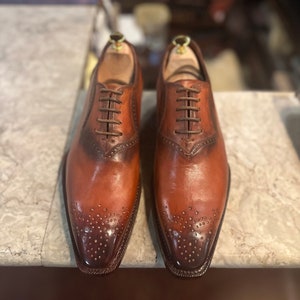 May include: A pair of brown leather oxford shoes with lace-up closures and a brogue design. The shoes have a pointed toe and a polished finish.