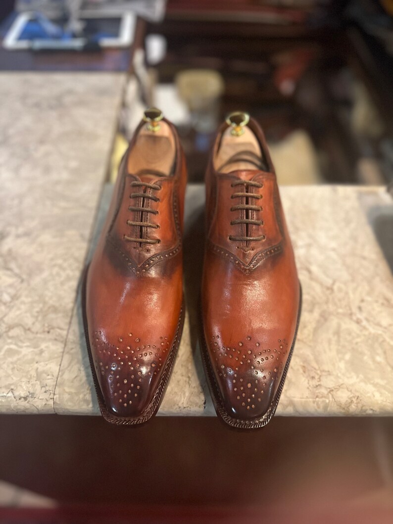 May include: A pair of brown leather oxford shoes with lace-up closures and a brogue design. The shoes have a pointed toe and a polished finish.