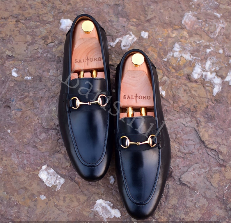 Op de afbeelding: Een paar zwarte leren loafers met gouden paardenbit accenten. De loafers hebben houten schoenspanners met de merknaam "Saltoro" erop gedrukt.