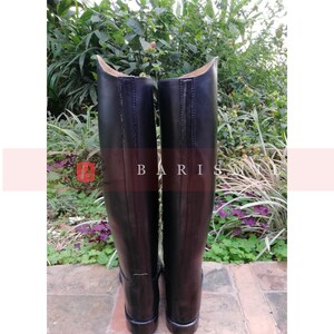 Puede incluir: Un par de botas de montar de cuero negro. Las botas tienen un acabado liso y pulido y una ca&ntilde;a alta. La parte superior de las botas es curva. La palabra "BARIS" est&aacute; impresa en las botas. Las botas est&aacute;n sobre un fondo de vegetaci&oacute;n.