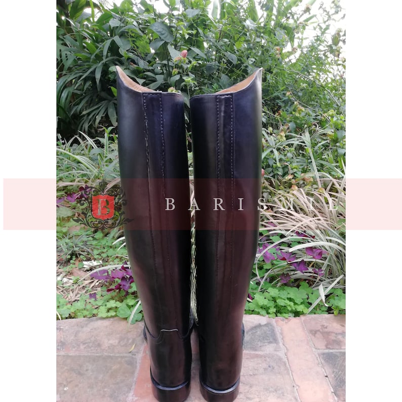 Puede incluir: Un par de botas de montar de cuero negro. Las botas tienen un acabado liso y pulido y una ca&ntilde;a alta. La parte superior de las botas es curva. La palabra "BARIS" est&aacute; impresa en las botas. Las botas est&aacute;n sobre un fondo de vegetaci&oacute;n.