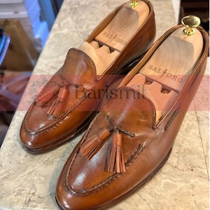 May include: A pair of brown leather tassel loafers with wooden shoe trees inside. The shoe trees are labeled "Saltoro".