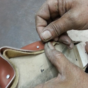 May include: A close-up of a person's hands working on a brown leather bag. The bag has a beige lining and is being stitched together with a needle and thread. The bag has metal rivets along the edges.