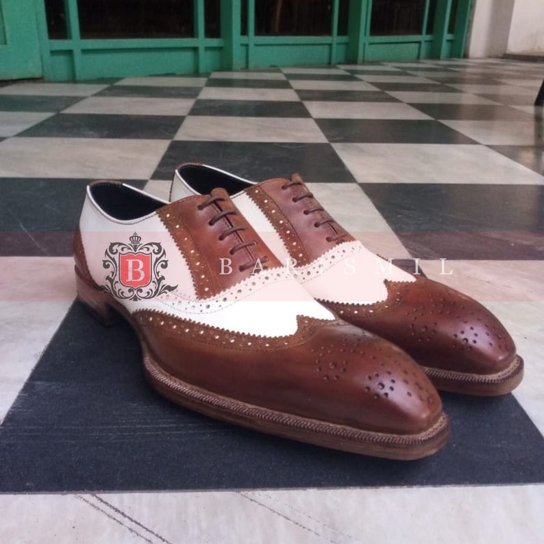 May include: A pair of brown and white leather brogue shoes with a wingtip design. The shoes have a lace-up closure and a white leather panel on the side. The shoes are on a chequered floor.