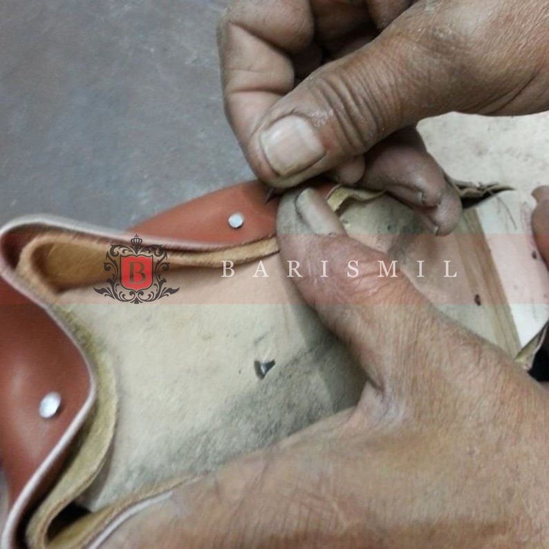 May include: Close-up of a person's hands sewing a brown leather piece with a needle and thread. The leather has a beige lining and the text "BARIS MIL" is visible on the piece.