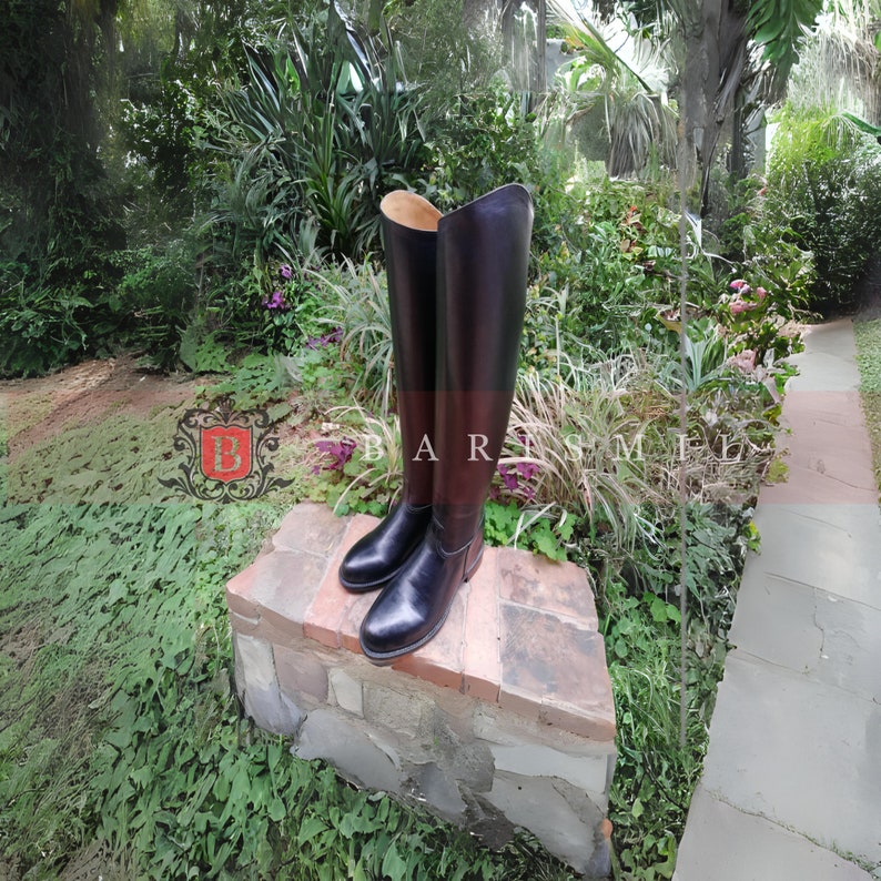 Puede incluir: Un par de botas altas de montar de cuero negro con un detalle marr&oacute;n en la pantorrilla. Las botas se exhiben al aire libre sobre una estructura de ladrillo y piedra, con el texto "BARNSMIL" visible.