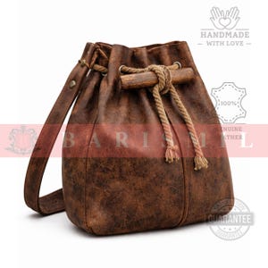 May include: Brown leather bucket bag with a drawstring closure and a shoulder strap. The bag features a wooden toggle and rope detailing. The bag has a distressed texture and is labeled "HANDMADE WITH LOVE" and "GENUINE LEATHER".