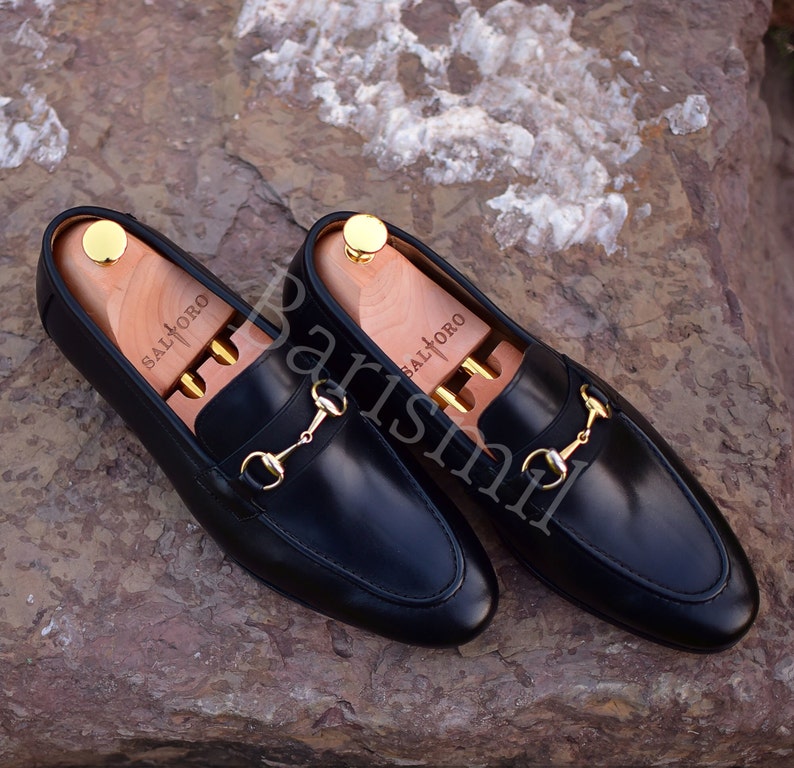 Op de afbeelding: Een paar zwarte leren loafers met gouden paardenbit accenten. De loafers hebben houten schoenspanners in de binnenkant.