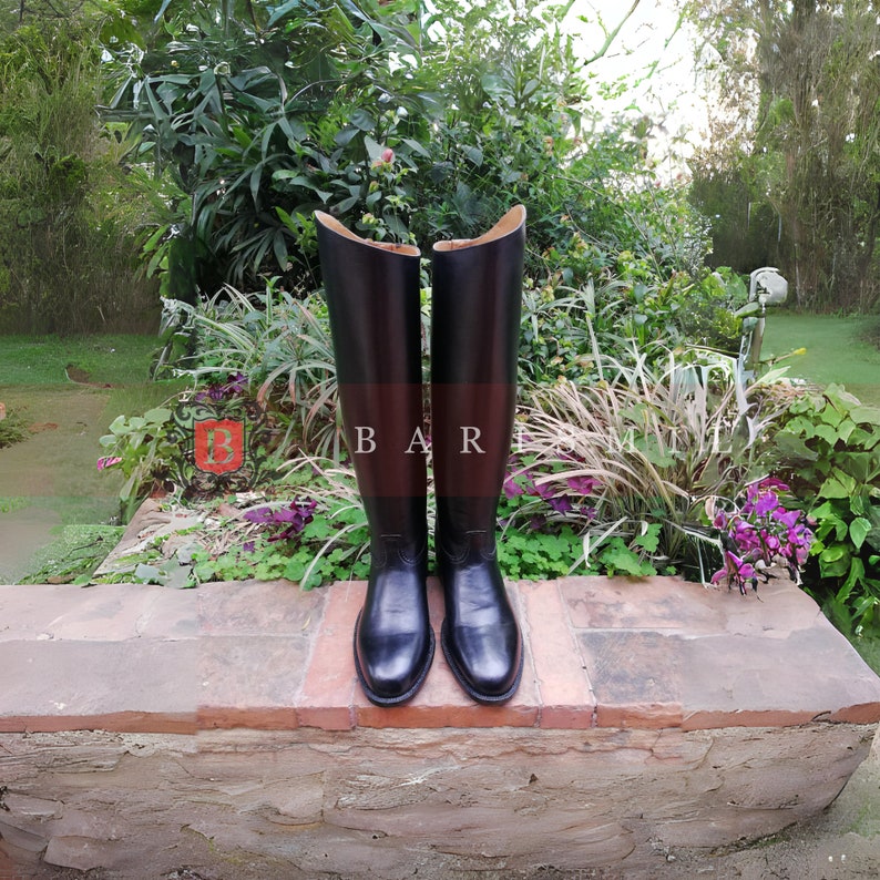 Puede incluir: Un par de botas de montar altas de cuero negro. Las botas se exhiben de pie, mostrando su dise&ntilde;o elegante y ca&ntilde;as altas. Se colocan sobre un borde de ladrillo, con un fondo de exuberante vegetaci&oacute;n.