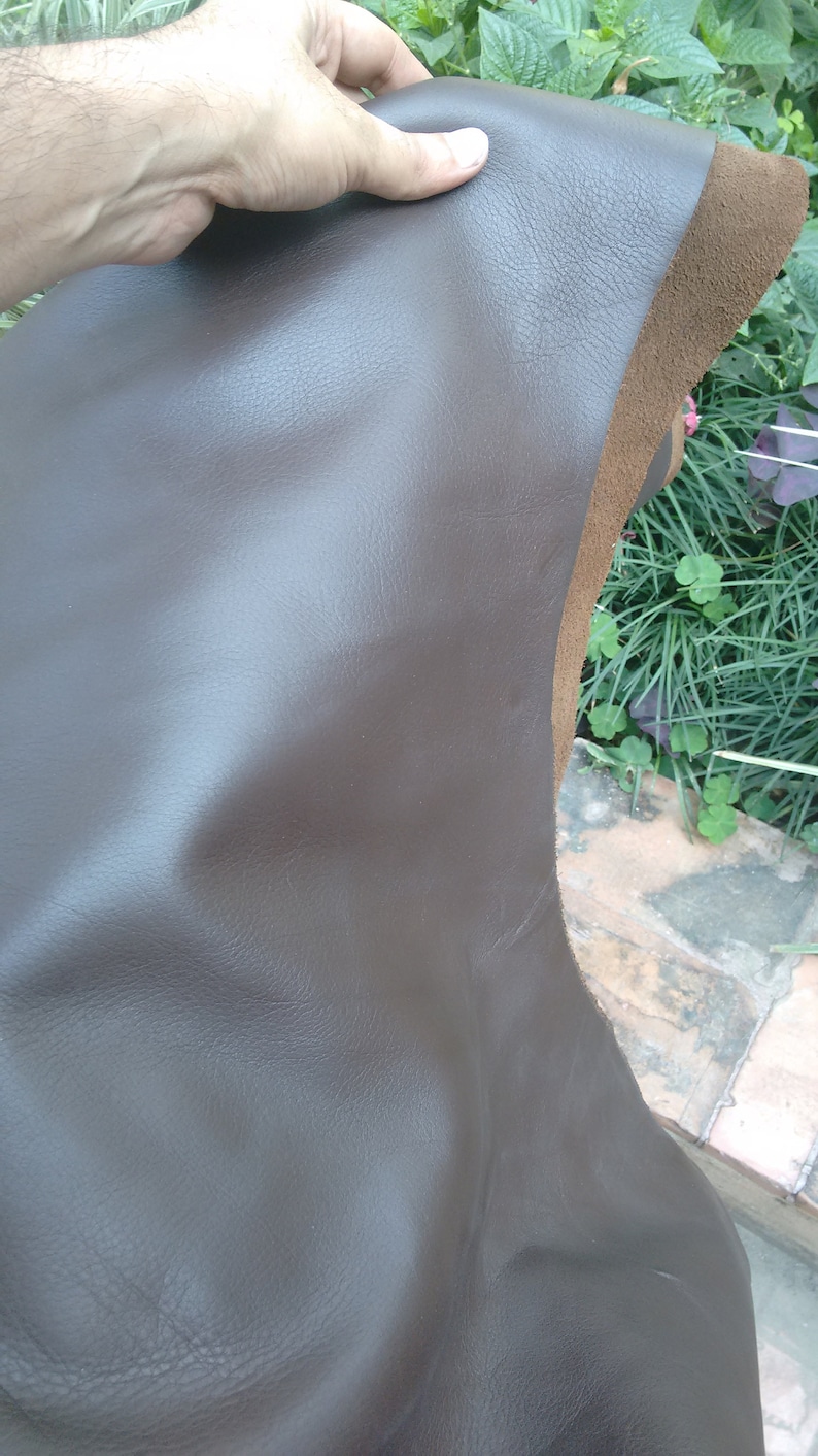 May include: A piece of brown leather with a smooth, textured surface. The leather is folded in half and held by a hand.