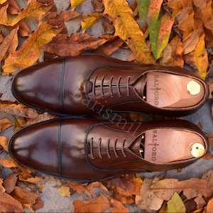 May include: A pair of brown leather oxford shoes with lace-up closures. The shoes are on a bed of autumn leaves. The brand name "SALTORO" is visible on the shoe.