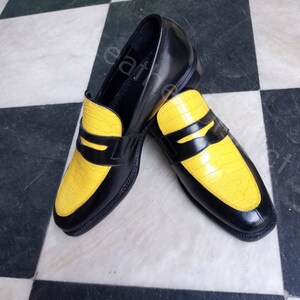 May include: A pair of black leather loafers with yellow crocodile-embossed leather accents. The loafers have a classic penny loafer design with a strap across the top.
