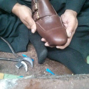 May include: A brown leather shoe with two buckles and a closed toe. The shoe is being held by a person's hands.