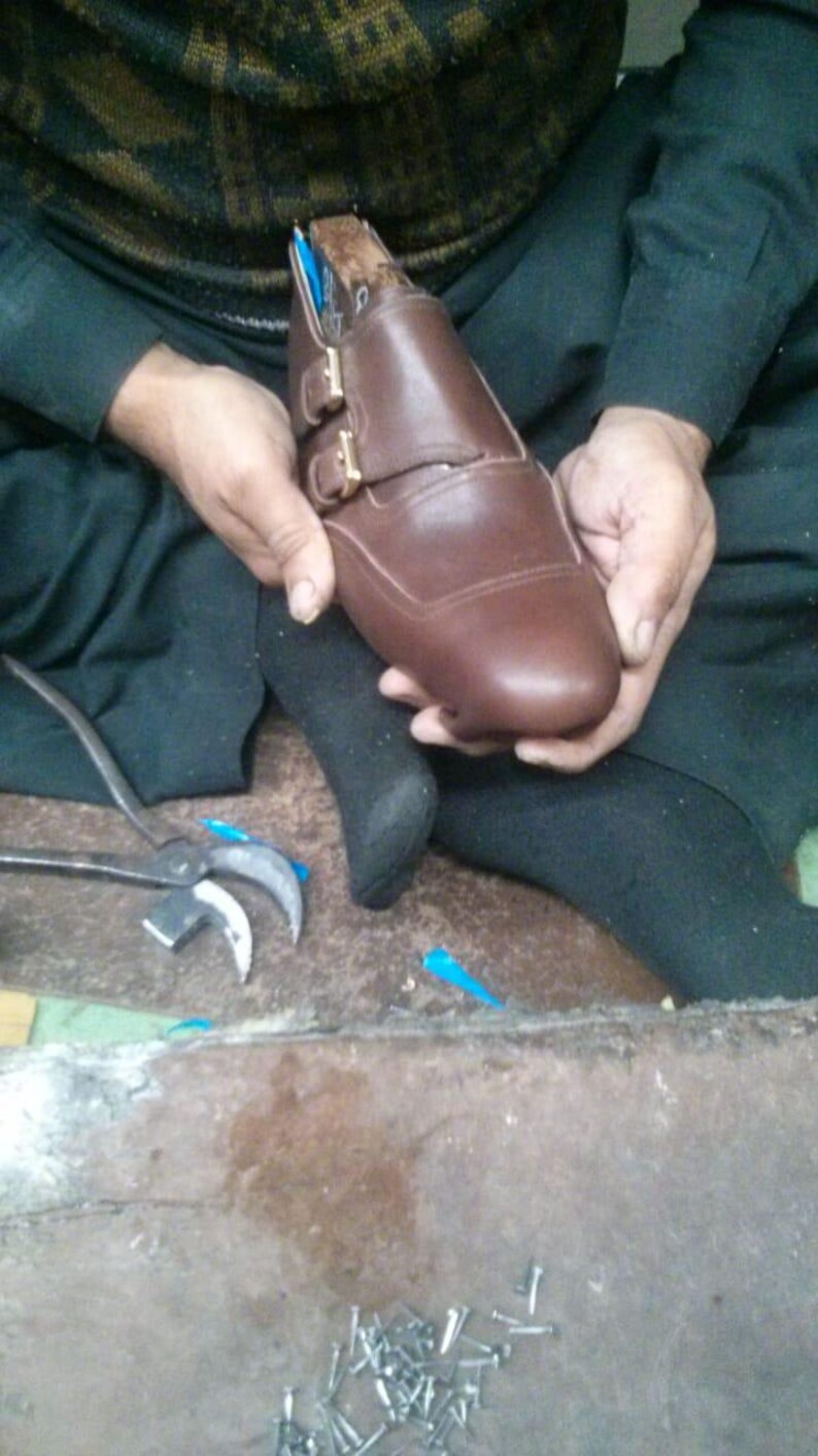 May include: A brown leather shoe with two buckles and a closed toe. The shoe is being held by a person's hands.