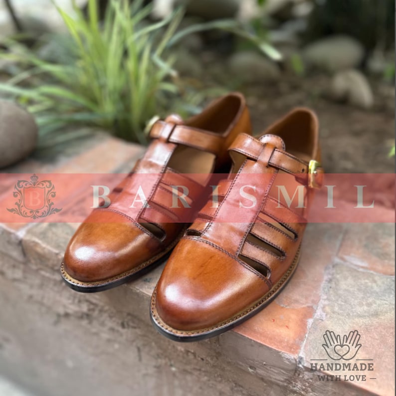 Puede incluir: Un par de sandalias de cuero marr&oacute;n con dise&ntilde;o de correa en T y cierres de hebilla. Las sandalias tienen un acabado pulido y est&aacute;n sobre una superficie de ladrillo. La imagen incluye el texto "BARISMIL" y "HANDMADE WITH LOVE".