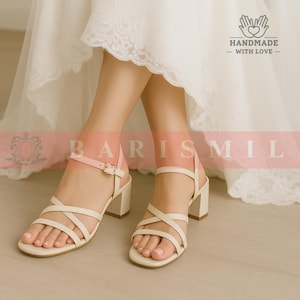 May include: Cream-colored strappy sandals with a block heel. The sandals feature multiple straps across the foot and ankle, with a buckle closure. The heels are a neutral color, and the sandals appear to be made of leather or a similar material. The sandals are part of a bridal collection.