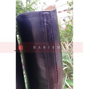 Puede incluir: Primer plano de una bota de montar de cuero negro. La bota tiene una superficie lisa y pulida con costuras visibles a lo largo de las costuras. El texto "BARISM" es visible en el lateral de la bota. El fondo est&aacute; borroso con vegetaci&oacute;n.