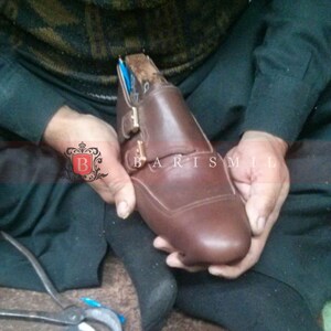 May include: A brown leather shoe with a buckle closure is being held by a person. The shoe is being worked on by a shoemaker.