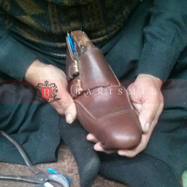 May include: A brown leather shoe with a buckle closure is being held by a person. The shoe is being worked on by a shoemaker.