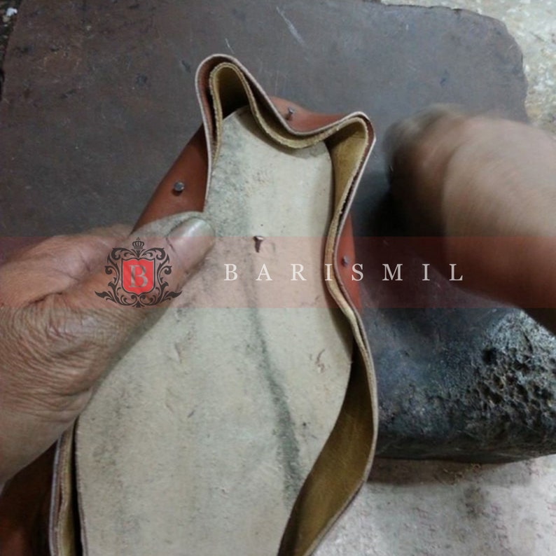May include: A close-up of a brown leather shoe sole being stitched together with a needle and thread. The sole is being held in place by a hand.