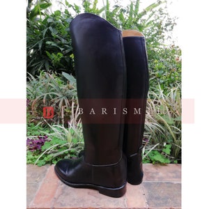 Puede incluir: Un par de botas de montar negras. Las botas est&aacute;n hechas de un material liso y brillante y tienen un dise&ntilde;o cl&aacute;sico. Las botas est&aacute;n de pie sobre una superficie de ladrillo, con vegetaci&oacute;n en el fondo. El texto "BARISM" es visible.
