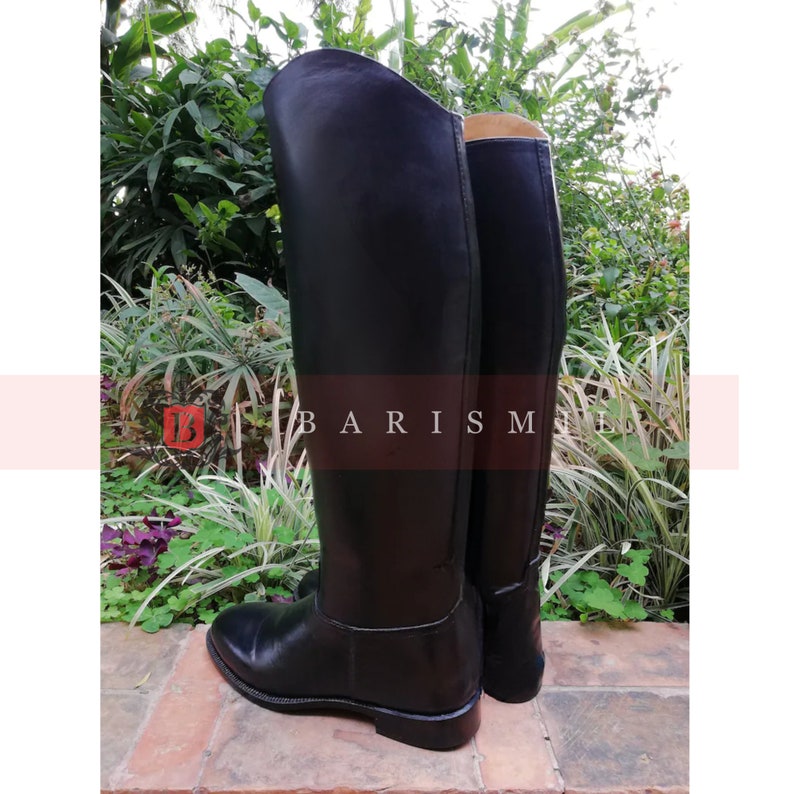 Puede incluir: Un par de botas de montar negras. Las botas est&aacute;n hechas de un material liso y brillante y tienen un dise&ntilde;o cl&aacute;sico. Las botas est&aacute;n de pie sobre una superficie de ladrillo, con vegetaci&oacute;n en el fondo. El texto "BARISM" es visible.