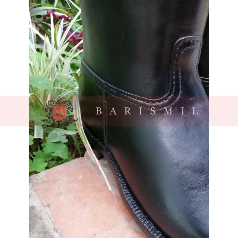Puede incluir: Primer plano de una bota de montar de cuero negro. La bota presenta un acabado liso y brillante y un dise&ntilde;o cosido a lo largo de las costuras. El texto "BARIS MIL" es visible en el lateral. La bota est&aacute; sobre un fondo de vegetaci&oacute;n.