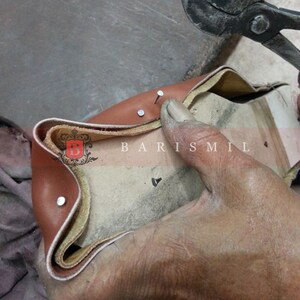 May include: Close-up of a brown leather shoe being stitched together with metal tacks. The shoe is being held by a hand and the tacks are being hammered in with a tool.