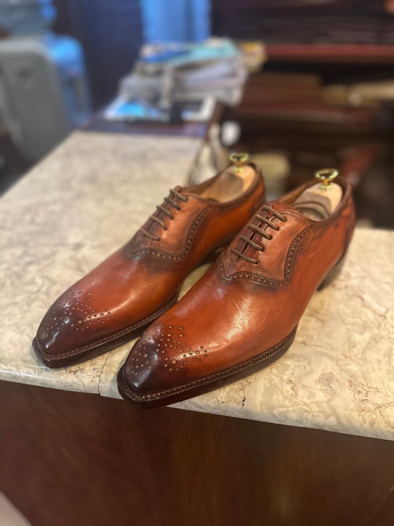 May include: A pair of brown leather oxford shoes with brogue detailing and lace-up closures. The shoes have a pointed toe and a polished finish.